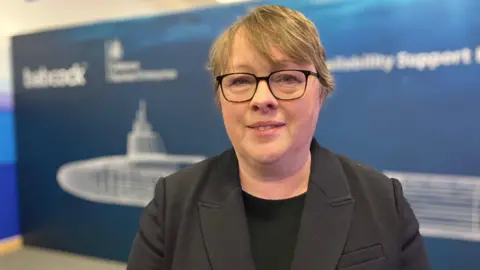 Maria Eagle, who has short blonde hair, glasses and is wearing a black blazer over a black top, smiles at the camera