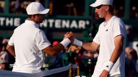 PA Media Two tennis players, both dressed all white, shake each others hands at the net on one of Wimbledon's grass courts. The crowd can be seen, blurred, in the background.