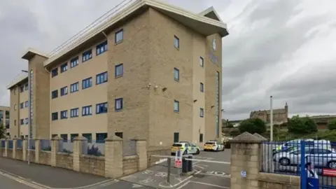 Google A beige four-storey building with a car park which is barriered off. Police vehicles are parked in the car park.