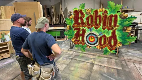 Three men wearing work clothes and navy T-shirts stand with their backs to the camera looking at a large sign reading "Robin Hood". One of the O's is a target with arrows in, and there are oak leaves around the words.