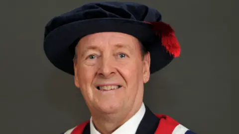 Nick Broady wearing a graduation cap and gown after receiving his honorary fellowship for his commitment to charity. He is smiling into the camera.