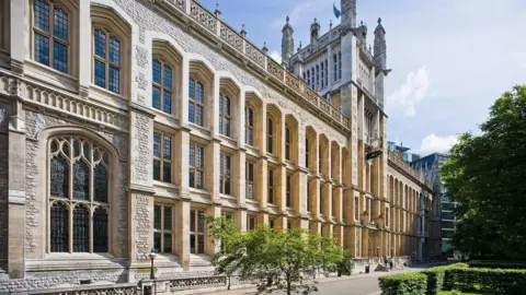 A university library which resembles a large chapel with a tower in the middle. There is a hedge and a tree on the right and a sapling in the foreground.