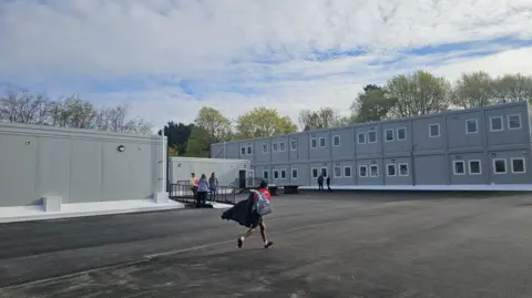 A grey two story building. A pupil with a red jumper is running towards the building carrying a backpack and a black coat.