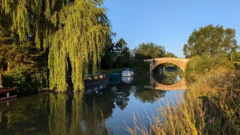 Morning sun on the river at Aston. There is a bridge over the river with a white boat and two narrow boats moored. A willow tree's branches overhang the water on the bank on the far side. On the right hand side the bank you can see long grass and purple flowers. The blue sky overhead is clear.