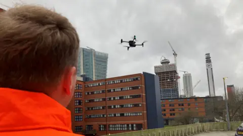 A drone in the sky in Birmingham. It is above buildings, including two which have cranes on. The BT tower is also on the skyline. The back of a man in an orange coat can also be seen. 