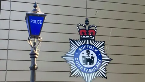 A Suffolk Constabulary badge on a wall with a blue police lamp-post in front of it. 