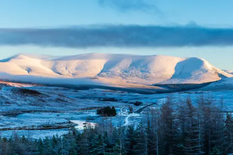 Angus Chisholm A distant mountain covered with snow and a valley below which is also dusted with white with trees in the foreground