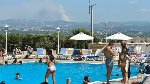 BBC / Goktay Koraltan People sunbathe and swim in a resort hotel as explosions smoke billows from a hillside in the distance after an Israeli strike 