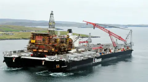 Heavy-lift vessel Pioneering Spirit carrying a large offshore oil and gas platform topside across calm coastal waters. A red lattice crane is positioned on the deck, with low-lying islands and shoreline visible in the background. The ship's hull displays the company name "Allseas."