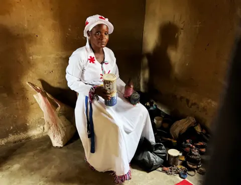 A woman in a white robe and hat with embroidered red flowers sits on chair in a dark room. Her shadow can be seen on one of the walls.