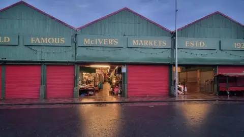 A row of three large sheds. Along the top of the sheds about red shuttered doors are signs reading: "World famous McIver's markets. Estd. 1921". Two of the doors are open and light spills on to a wet road.