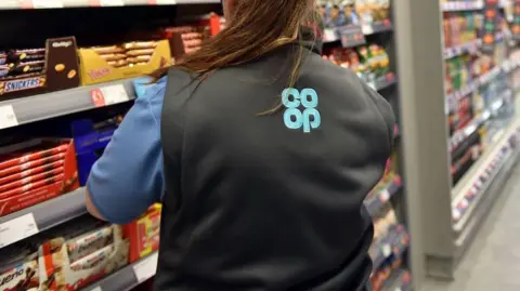 A woman with a long brown ponytail wears a black gillet with a blue co-op logo while looking at a shelf full of chocolates in the supermarket.