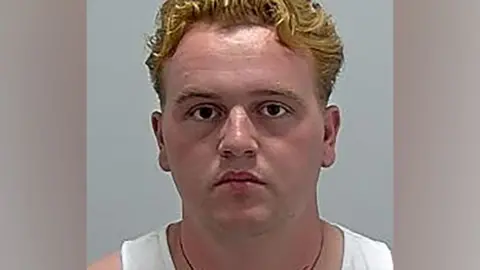A police custody mugshot of Johnny Ward. He is wearing a white vest and a necklace.,