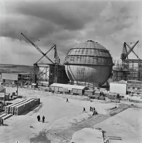 Getty Images In a black and white image, the sphere looks like a big metal ball. There are large cranes, scaffolding and temporary buildings nearby. Workers walk across a large space in front of the sphere. 