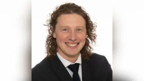 North Yorkshire Council A man with long curly hair wearing a dark suit jacket, white shirt and tie, posed against a plain white background in a formal portrait style.