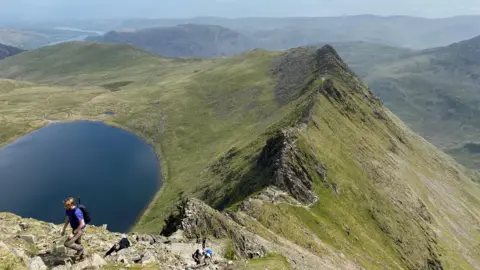Duncan Leatherdale A walker in a blue shirt nears the top of a steep rocky hill. Beyond him is a craggy line of rocks with steep sides either side sloping down. The slope on the left ends in a large blue tarn. There are other hills beyond.