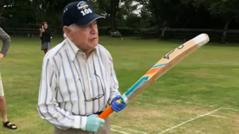 Family Geoffrey Doyle stands with a cricket bat in his hand. He is wearing gloves and a cap. 