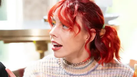 A woman with red hair, Jessie, is talking to someone sitting next to her, who is not visible. She is inside but the setting is not clear as the background is out of focus.