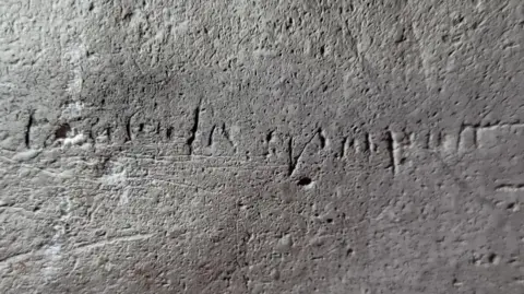 A stone grey wall. Etched on it are letters, barely distinguishable 