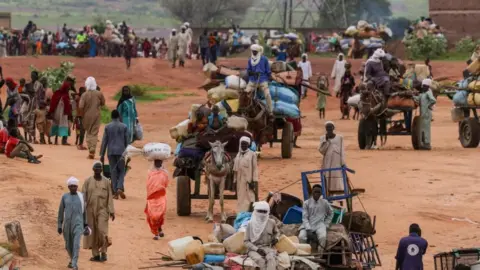 Reuters Um grupo de pessoas andando por um caminho em Darfur, alguns carregando itens na cabeça ou nas mãos. Um grupo de pessoas se senta em vários carrinhos diferentes, liderados por burros, com itens empilhados no carrinho.