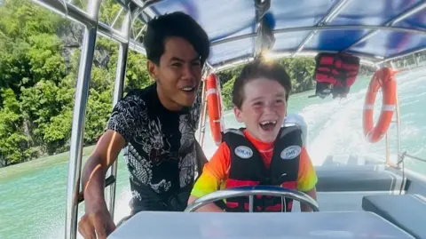 Alfie Kiely Alfie laughing while steering a boat. The sea and trees visible in the background