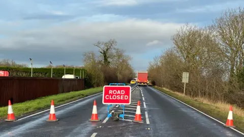 The scene of a crash on the A15. Five orange and white cones have been spread across the carriageway and a red sign which says "Road Closed" stands in the centre of the tarmac. The road is lined by a brown wooden fence on the left and trees on the right. A police car and a lorry are visible in the distance.