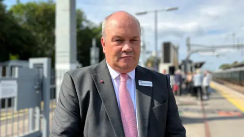 Mark Hopwood looks toward the camera on a station platform wearing a grey suit, white shirt and pink tie with a serious facial expression