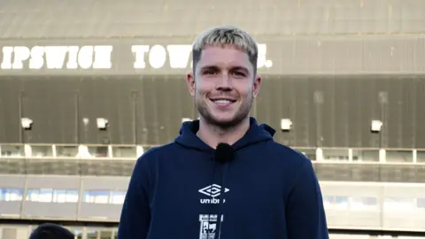 Tom Williams/BBC man wearing a hoodie standing in front of a stadium with 'IPSWICH TOWN' signage on the side in white text.
