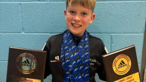 Sharon Gill Theo wearing lots of medals around his neck and holding two trophies, one in each hand. He is smiling at the camera