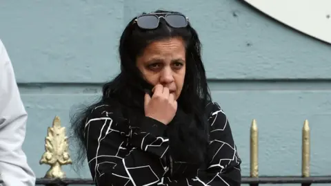 RTE A woman with long black hair with her hand up to her mouth. She has a pair of sunglasses pushed up onto her head and is standing before railings.
