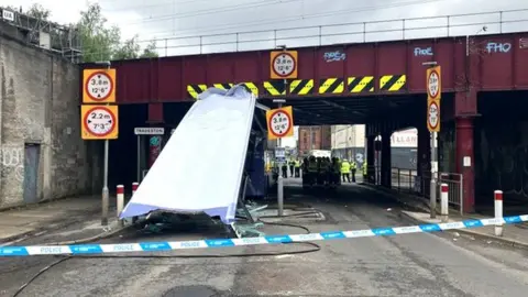 A bus roof sits at a 45 degree angle under a bridge. Police tape can be seen in the foreground. 