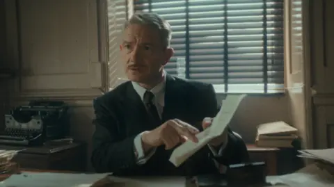 Justin Downing/Netflix Martin Freeman with a moustache in a suit. He is sitting at a desk and holding out a piece of paper which he is pointing at.