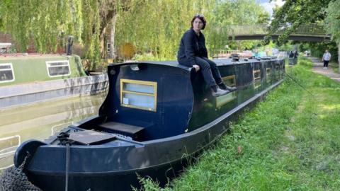 BBC reporter Clodagh Stenson sits on the roof of her narrowboat on the Oxford canal with a grass verge next to the tow path