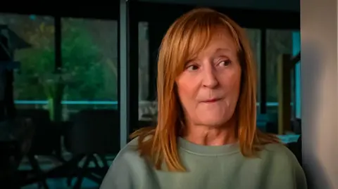 Diane is at the door of her home, which has big windows looking out onto trees. she is wearing a green cotton jumper and has red shoulder length hair. He mouth is closed and she has a look of concern on her face.
