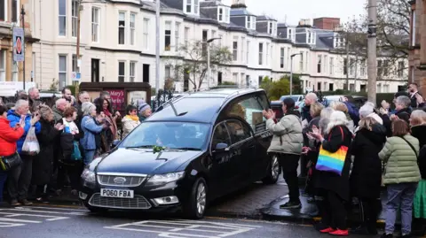 PA Media A achromatic  hearse successful  the mediate  of a roadworthy  with  a tiny  assemblage  of radical   wearing lukewarm  looking jackets and carrying bags clapping their hands.