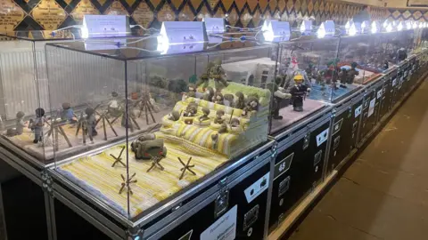 Two lines of clear boxes can be seen, with white labels on top to explain the scenes in each. There are various scenes depicting World War Two battles - with soldiers, tanks, beaches and trenches all depicted in wool.