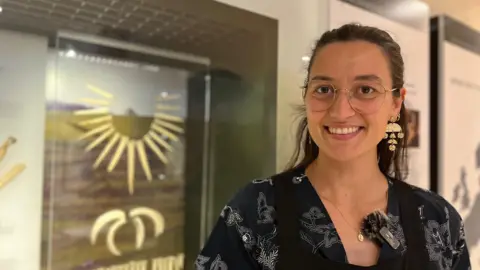 BBC Amber smiles at the camera. She stands in front of a blurred cabinet with items in.
She wears glasses and black floral top.