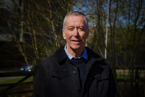 David King has a wry smile towards the camera. He wears a blue Oxford shirt, a blue ribbed woollen jumper and a black denim jacket