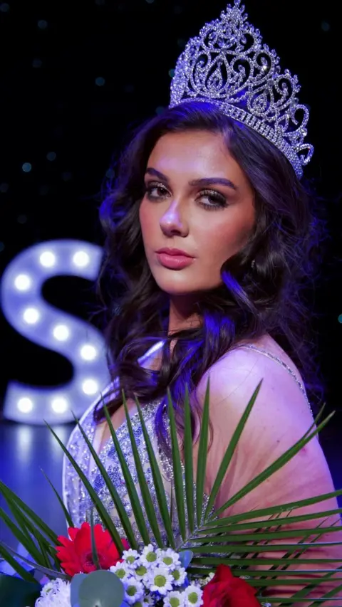 Miss Wales/Danielle Latimer A woman with long dark wavy hair, wearing a crown and a sparkly gown, holding a bouquet of flowers