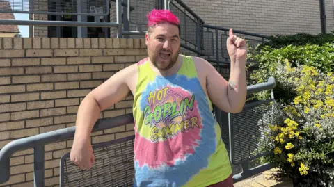 Aaron Weight stands in front of the BBC Radio Suffolk building, he is dressed in a blue, pink and yellow tank top. His hair is bright pink and he is smiling while pointing up to the sky.