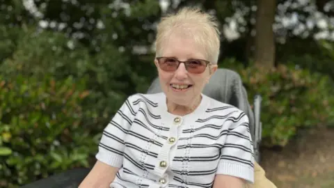 A woman with short blonde hair is smiling and sat in a chair outside with shrubbery and trees behind her. She is wearing a white and black stripey top and tinted glasses.