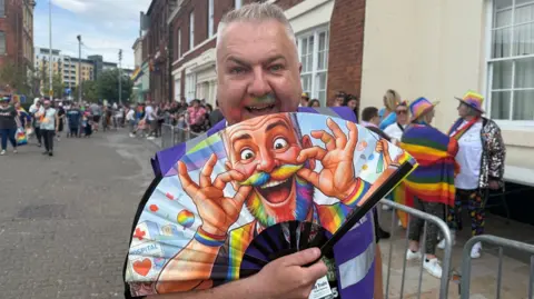 Train holds a stretched out fan with a picture of himself on it. It depicts him with a rainbow beard and mustache. Other people are standing in the street behind him.