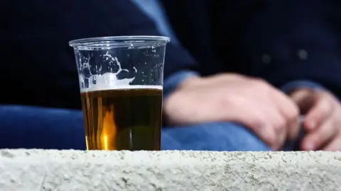 A pint of beer in a plastic cup sitting on a wall. A person can be seen sitting in the background.