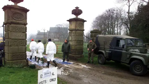 A soldier guards the entrance to a farm as three people in white hazmat suits walk onto the premesis.