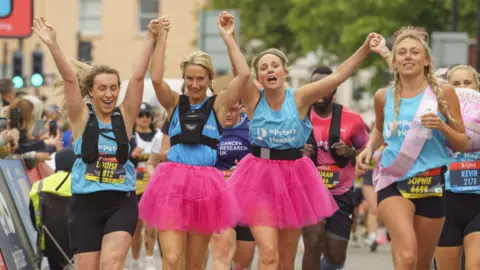 Three young women smile and link hands as they run in the Bristol Half Marathon. Two of them are wearing pink skirts below their running gear