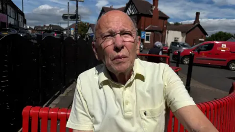 Gemma Dillon A man wearing a yellow t-shirt is sitting on a red bench..
He has a bald head and is wearing glasses