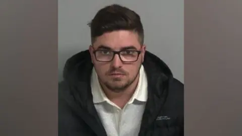 Bedfordshire Police A mugshot of Elliot Wright wearing a black coat, gray collared sweater and black-rimmed glasses. He has brown hair and a stubble beard. He stares into the camera with a serious expression.