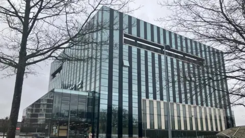 The outside of a glass-panelled building. It has a number of trees in front of it. 