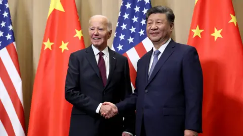 Reuters U.S. President Joe Biden and Chinese President Xi Jinping shaking hands in front of a backdrop displaying the flags of the United States and China. Biden is dressed in a dark suit with a white shirt and a burgundy tie, while Xi wears a navy suit paired with a white shirt and a blue tie.