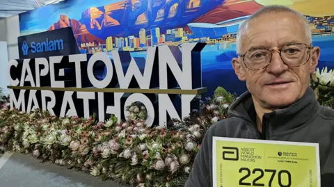 Nick Price A man with short grey hair and glasses holding up a yellow sign with the number 2270 in front of a large sign which reads Cape Town Marathon